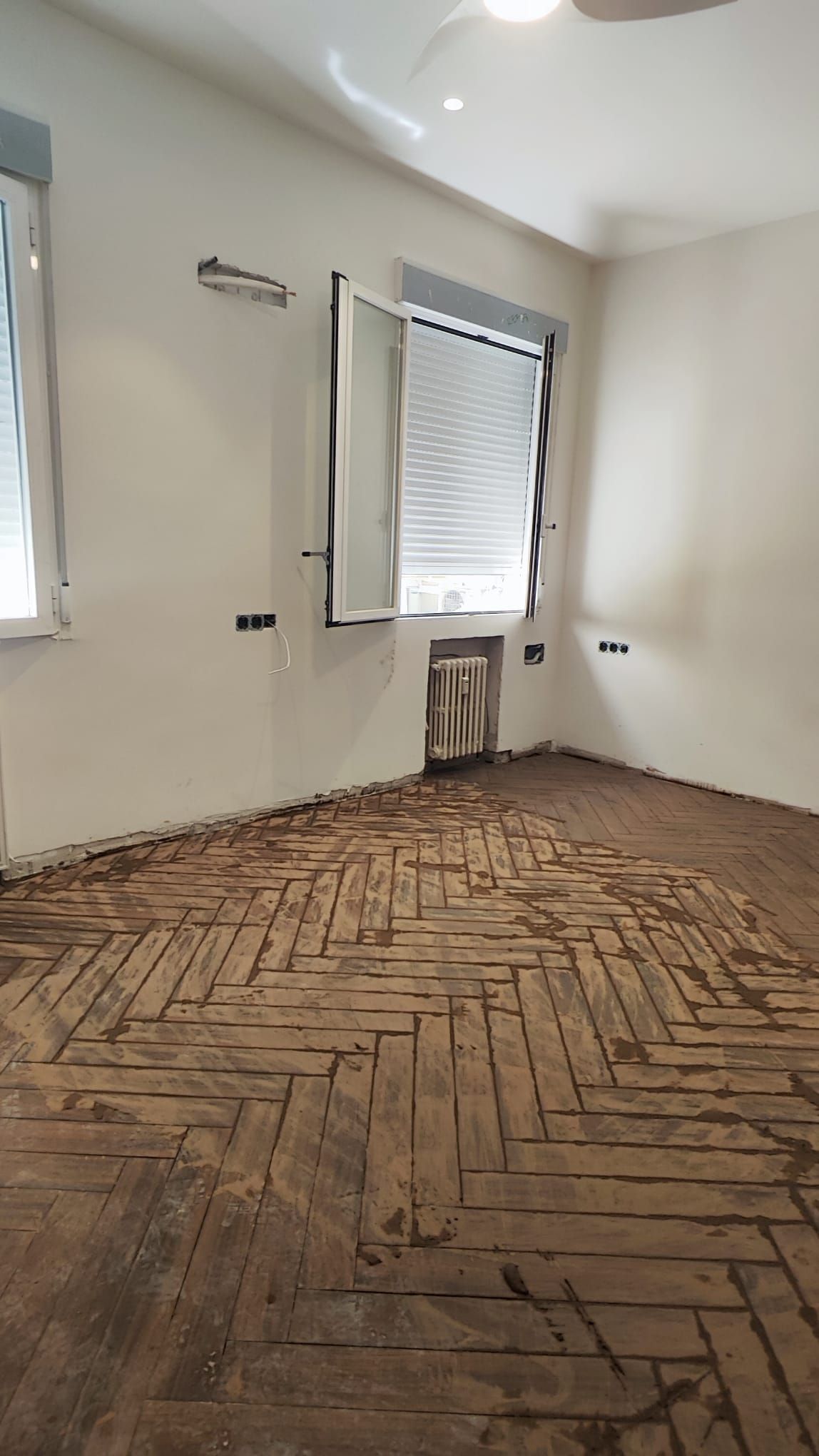 Salón en obra con el suelo de parqué en espiga y la ventana con persiana